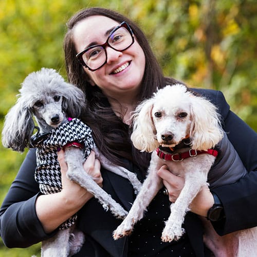 A person wearing glasses, smiling like a proud veterinarian, holds two small poodles. One poodle flaunts a black and white patterned coat, while the other sports a gray vest. The background is a blur of lush greenery.