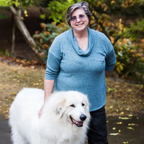 A smiling woman wearing glasses and a blue sweater stands outdoors, her hand gently resting on a large, fluffy white dog. As a veterinarian, she appreciates moments like these amid nature's green foliage and the shade of a nearby tree.