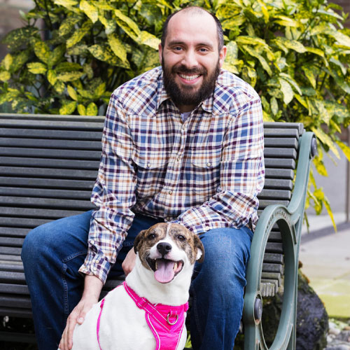 A man in a plaid shirt sits on a bench with his smiling, brindle and white dog, who wears a pink harness. The dog, healthy and full of energy thanks to regular visits to the vet, enjoys the lush green foliage in the background.